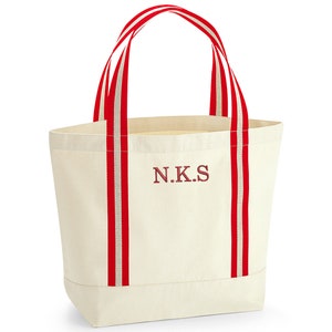 May include: A white canvas tote bag with red and white striped handles. The bag has the initials "N.K.S." embroidered on the front in red.