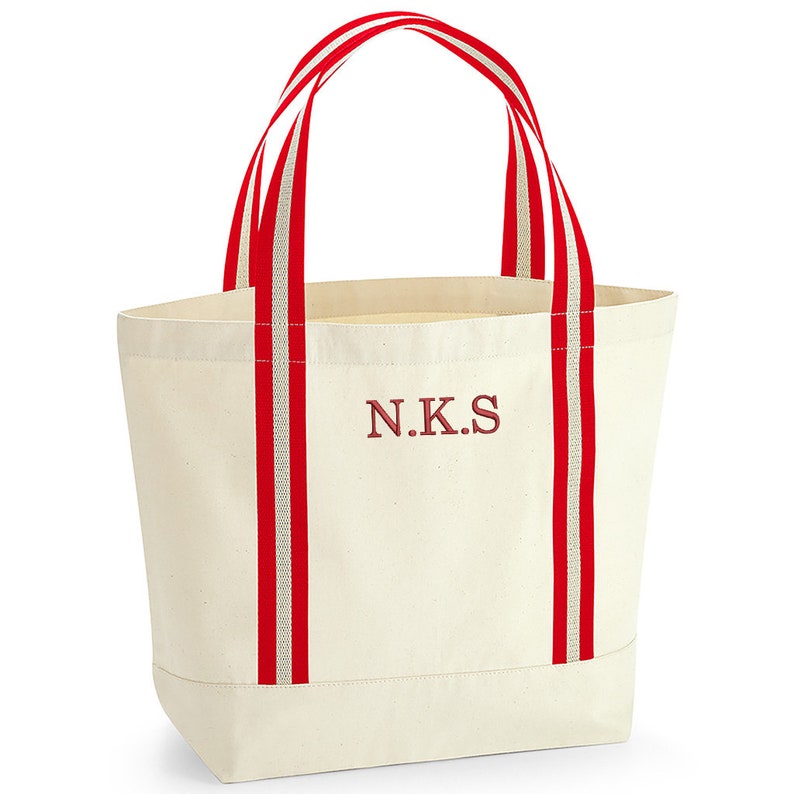 May include: A white canvas tote bag with red and white striped handles. The bag has the initials "N.K.S." embroidered on the front in red.