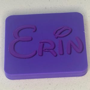 May include: A rectangular purple silicone mould featuring the name Erin embossed in a stylised script font on the surface. The mould has a smooth, flat base and raised edges, with the letters clearly defined in a recessed, darker purple tone.
