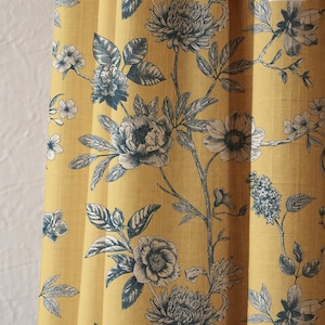 May include: A close-up of a yellow curtain panel with a floral design. The fabric displays a detailed botanical print with blue and white flowers and leaves. The design creates a classic, elegant aesthetic, suitable for home decor.
