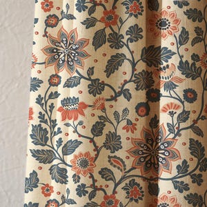 May include: Cream-colored fabric with a repeating floral pattern. The design features large, stylized flowers in shades of coral and navy blue, with intricate details and leafy vines. The background is a neutral beige.