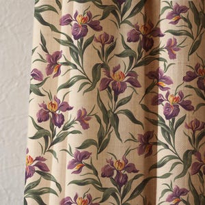 May include: A cream-coloured fabric with a repeating floral pattern. The design features purple iris flowers with yellow and orange centres, green leaves, and stems. The fabric has a textured appearance, possibly linen.
