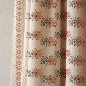 K&ouml;nnte beinhalten: Beigefarbener Stoff mit einem sich wiederholenden Paisley-Muster in Orange, Gr&uuml;n und Braun. Ein dekorativer Rand mit floralen und wellenf&ouml;rmigen Designs verl&auml;uft entlang der linken Kante. Der Stoff hat ein strukturiertes Aussehen.