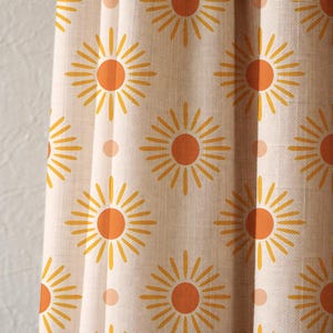 May include: Cream-coloured fabric featuring a repeating pattern of orange and yellow sunburst designs. Each sunburst has a central orange circle with yellow rays. The fabric also has small, pale orange polka dots.