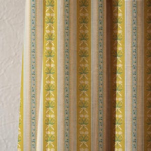 May include: Close-up of a fabric with vertical stripes in shades of yellow, blue, and white. The design includes floral and ribbon patterns. The fabric appears to be a curtain or upholstery material.