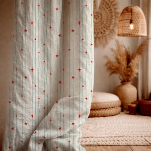 May include: White curtain with vertical light blue stripes and scattered red star patterns. The curtain is made of a light fabric and is partially draped, revealing a cozy interior with natural textures and warm lighting.