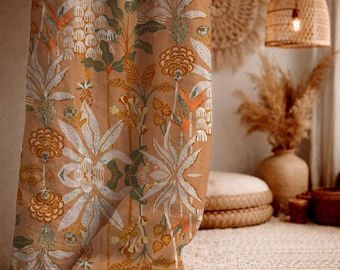 May include: A tan curtain with a repeating floral pattern in white, green, and orange. The design features stylized flowers and foliage. The curtain is hanging in front of a neutral-toned background, suggesting a bohemian or natural aesthetic.