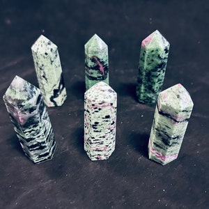 May include: Six green, black, and pink gemstone points, also known as green uvarovite, are arranged in a circle on a black surface.