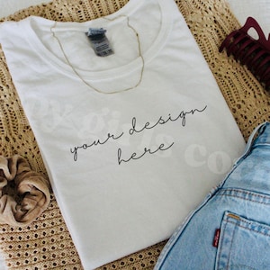 May include: A white t-shirt with the text "your design here" in black script. A gold necklace rests on the shirt, which is paired with blue denim jeans and a brown scrunchie. A burgundy hair clip is also visible.