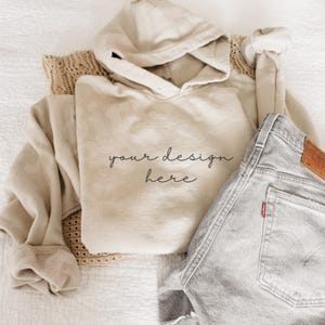 May include: A beige hoodie with the text "your design here" is folded next to a pair of gray denim shorts. A tan knit sweater is partially visible. The items are arranged on a white, textured surface.