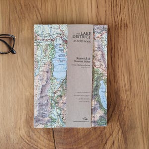 May include: A5 notebook featuring a map of the Lake District, Keswick & Derwent Water. The notebook has a brown paper band with text. A pair of black glasses and a silver pen are on a wooden surface.