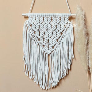 Könnte beinhalten: Weißer Makramee-Wandbehang mit geometrischem Muster und Fransen. Der Behang ist aus Baumwollschnur gefertigt und an einer Holzleiste befestigt.