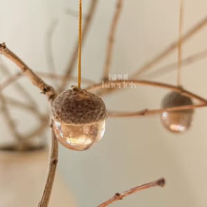 Könnte beinhalten: Nahaufnahme von dekorativen Eichelornamenten, die an dünnen Zweigen hängen. Die Eicheln haben eine braune Kappe und einen klaren, abgerundeten Boden. Die Zweige sind hellbraun, mit einer weißen Vase im Hintergrund.