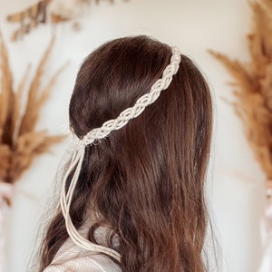 May include: A white braided headband worn on a person's head. The headband is made of a thick rope and has a simple design.
