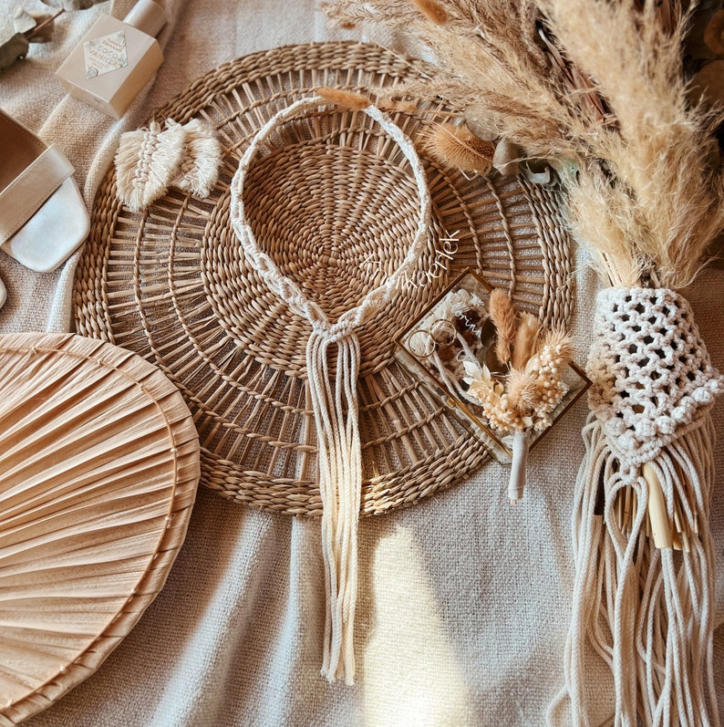 May include: A white macrame necklace with a tassel hangs on a woven wicker mat. The necklace is made of cotton cord and has a simple design. The mat is round and has a natural brown color.