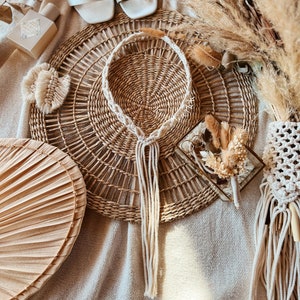 May include: A white macrame cord necklace with a long tassel hangs on a woven straw mat. The mat is surrounded by dried pampas grass and other natural elements.