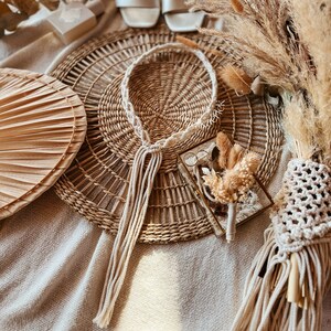 May include: A white macrame cord necklace with tassels hangs on a woven wicker mat. A small glass box with dried flowers and a pair of glasses sits on the mat.