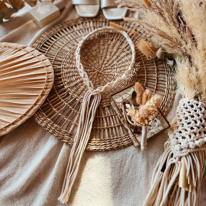May include: A white macrame cord necklace with tassels hangs on a woven wicker mat. A small glass box with dried flowers and a pair of glasses sits on the mat.
