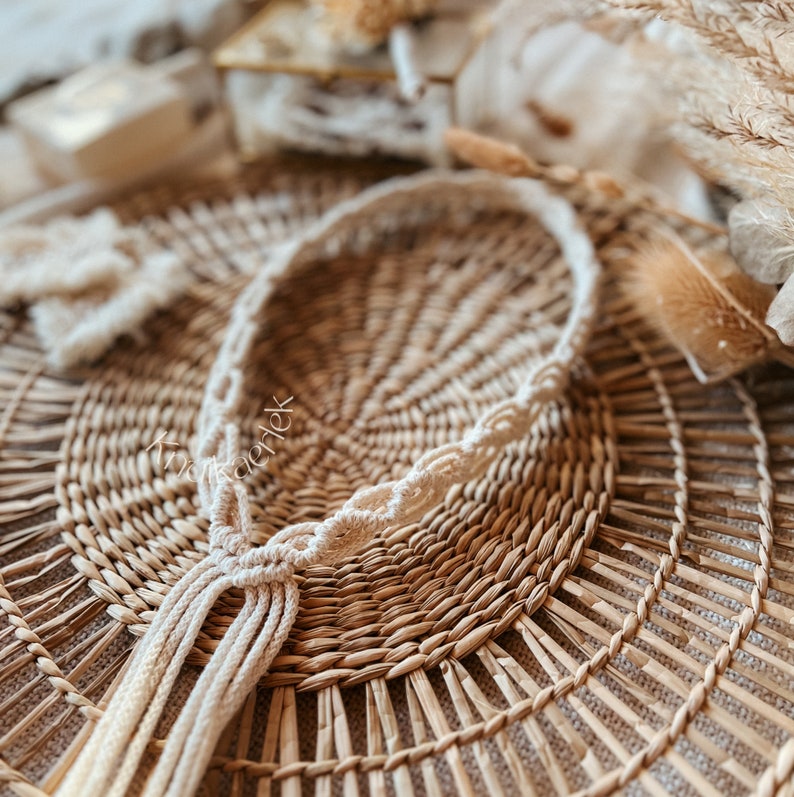 May include: A white macrame plant hanger with a knotted tassel, hanging on a woven wicker surface.