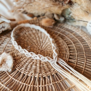 May include: A white macrame cord necklace with a tassel on a woven straw background.