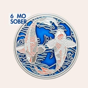 Può includere: Una moneta di sobrietà in metallo circolare con due pesci koi che nuotano in un motivo d'acqua blu. I pesci sono bianchi con accenti arancioni e squame dettagliate. Il testo 6 MO SOBER è stampato in blu sul lato sinistro della moneta.