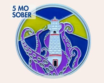 May include: A round metal token featuring a white lighthouse with red accents surrounded by a purple octopus with visible suckers. The background shows a yellow sky and blue sea. The text 5 MO SOBER is printed in blue on the upper left side.