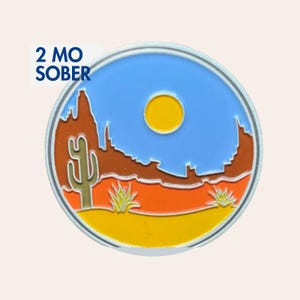Può includere: Una spilla rotonda in metallo raffigurante un paesaggio desertico con un sole giallo, cielo blu, formazioni rocciose marroni, un cactus saguaro verde e terreno arancione e giallo. Il testo 2 MO SOBER è stampato in blu scuro in alto a sinistra.