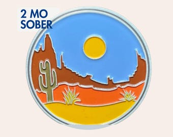May include: A round metal pin featuring a desert landscape with a yellow sun, blue sky, brown rock formations, a green saguaro cactus, and orange and yellow ground. The text 2 MO SOBER is printed in dark blue in the upper left corner of the pin.
