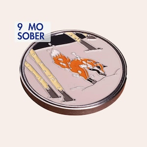 Può includere: Una moneta rotonda in metallo per la sobrietà con sfondo rosa chiaro e una volpe arancione stilizzata che salta sopra due tronchi di betulla gialli. Il testo 9 MO SOBER è stampato in blu scuro nell'angolo in alto a sinistra della medaglia.