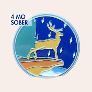 Può includere: Un gettone di sobrietà rotondo in metallo raffigurante un cervo dorato in piedi su una scogliera contro un cielo notturno blu scuro stellato. Il design include una sezione verde acqua a sinistra e terreno a strati arancione e giallo. Testo 4 MO SOBER.