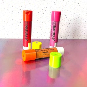 Könnte beinhalten: Lippenbalsam in roten, rosa- und orangenfarbenen Tuben mit weißen Basen. Die Tuben sind mit "BEENATURAL 100% NATURAL LIP BALM" und "MANGO" beschriftet. Zwei gelbe und grüne Kappen sind ebenfalls sichtbar. Der Lippenbalsam ist 4,2 g.