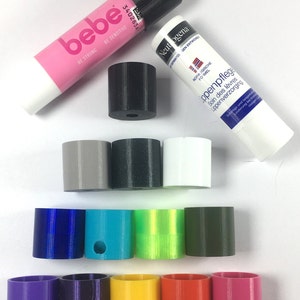 Könnte beinhalten: Eine Sammlung von 11 verschiedenen farbigen Plastikkappen für Lippenbalsam-Tuben. Die Kappen sind in drei Reihen angeordnet. Die obere Reihe hat drei Kappen: grau, schwarz und weiß. Die mittlere Reihe hat vier Kappen: blau, türkis, grün und dunkelgrün. Die untere Reihe hat vier Kappen: lila, gelb, orange und rosa.