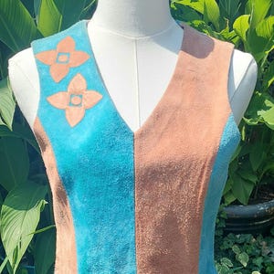 May include: A sleeveless suede dress featuring alternating vertical panels of tan and bright turquoise. The bodice has a V-neckline and two tan flower-shaped appliqués on the right shoulder, while the skirt features two turquoise flower-shaped appliqués.