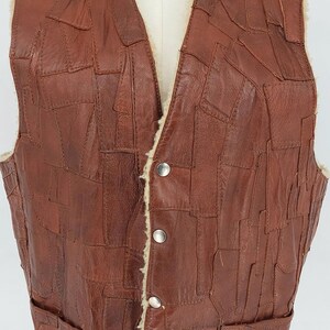 May include: Brown leather vest with a patchwork design. The vest has a V-neck, two front pockets, and a cream-colored lining. It fastens with three silver buttons. The vest is displayed on a mannequin.