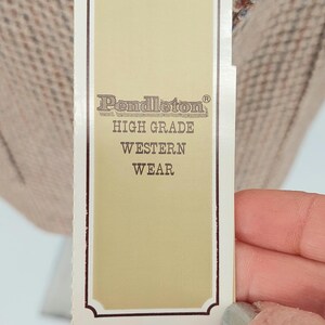 Peut inclure: Une &eacute;tiquette de produit Pendleton, avec le nom de la marque et le texte "HIGH GRADE WESTERN WEAR". L'&eacute;tiquette est de couleur cr&egrave;me avec une bordure fonc&eacute;e et est attach&eacute;e &agrave; un tissu &agrave; motifs.
