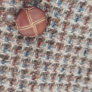 Peut inclure: Gros plan d'un bouton rond marron avec un motif en point de croix. Le bouton est pos&eacute; sur un tissu tweed textur&eacute; aux tons marron, bleu et rouge.