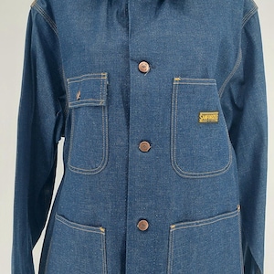 May include: A blue denim jacket with four front pockets and button closures. The jacket has a collared neckline and the text "SANFORIZED" is visible on one of the pockets. The jacket is displayed on a mannequin.