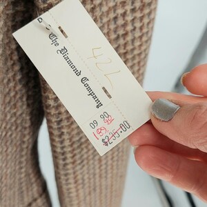 Peut inclure: Gros plan sur une veste en tweed dans des tons marron, beige et bleu. Le tissu pr&eacute;sente un motif pied-de-poule classique. Une &eacute;tiquette de "The Diamond Company" est visible.