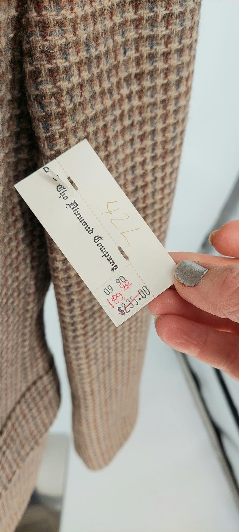 Peut inclure: Gros plan sur une veste en tweed dans des tons marron, beige et bleu. Le tissu pr&eacute;sente un motif pied-de-poule classique. Une &eacute;tiquette de "The Diamond Company" est visible.