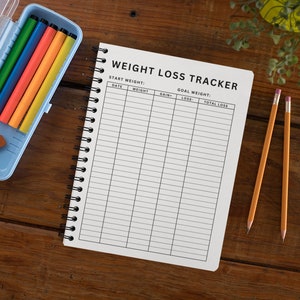 May include: A weight loss tracker journal with a black and white design. The journal has a spiral binding and is open to a page with the title "Weight Loss Tracker". The page has columns for date, weight, gain, loss, and total loss. There are also colored pencils and a pencil on the table.