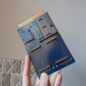 Könnte beinhalten: Eine blaue Leiterplatte mit goldenen Anschlüssen und mehreren integrierten Schaltkreisen. Die Platine trägt den Text "T.D.T." und weitere Markierungen. Sie wird in einer Hand vor einem neutralen Hintergrund gehalten.