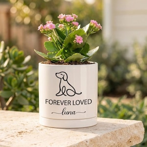May include: A white ceramic cylindrical planter containing a small flowering plant with pink blossoms. The front of the planter features a black line art drawing of a sitting dog above the text FOREVER LOVED and the name Luna in script.