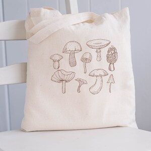 May include: A white canvas tote bag with a brown line drawing of various mushrooms. The mushrooms are drawn in a simple, realistic style.