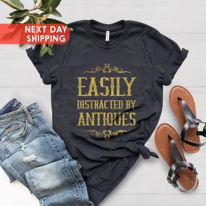 May include: A black t-shirt with gold text that reads 'Easily Distracted by Antiques'. The shirt is folded and laid flat on a white wooden surface. There are blue jeans and brown sandals next to the shirt.