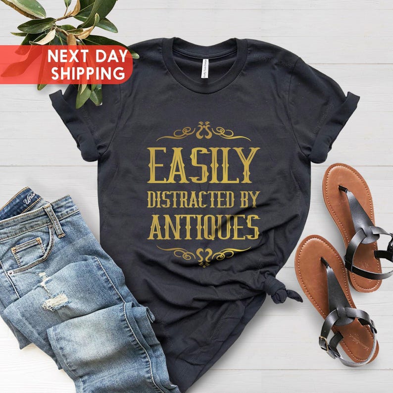May include: A black t-shirt with gold text that reads 'Easily Distracted by Antiques'. The shirt is folded and laid flat on a white wooden surface. There are blue jeans and brown sandals next to the shirt.