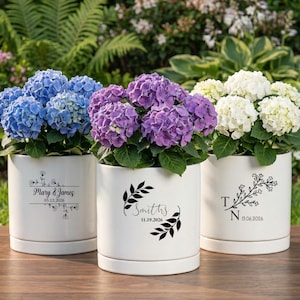 May include: Three white cylindrical ceramic planters, each featuring a black botanical design and personalized text. The left planter displays 'Mary & James 05.12.2026', the center 'Smiths 11.19.2026', and the right 'T N 13.06.2026', filled with blue, purple, and white hydrangeas.