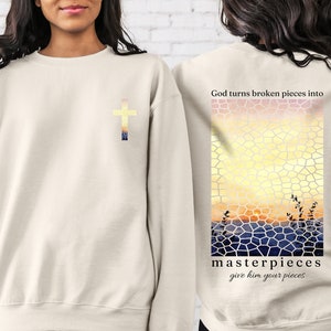 May include: Beige sweatshirt with a cross on the front and a stained glass sunset design on the back. The text "God turns broken pieces into masterpieces give him your pieces" is printed on the back.