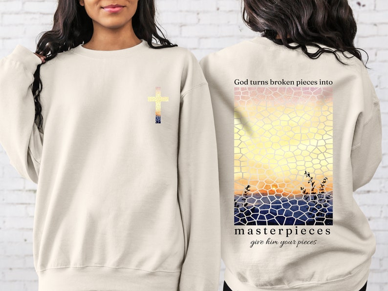 May include: Beige sweatshirt with a cross on the front and a stained glass sunset design on the back. The text "God turns broken pieces into masterpieces give him your pieces" is printed on the back.