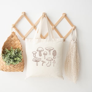 May include: A white tote bag with a brown line drawing of various mushrooms. The bag is hanging on a wooden rack with a woven basket and a mesh bag.