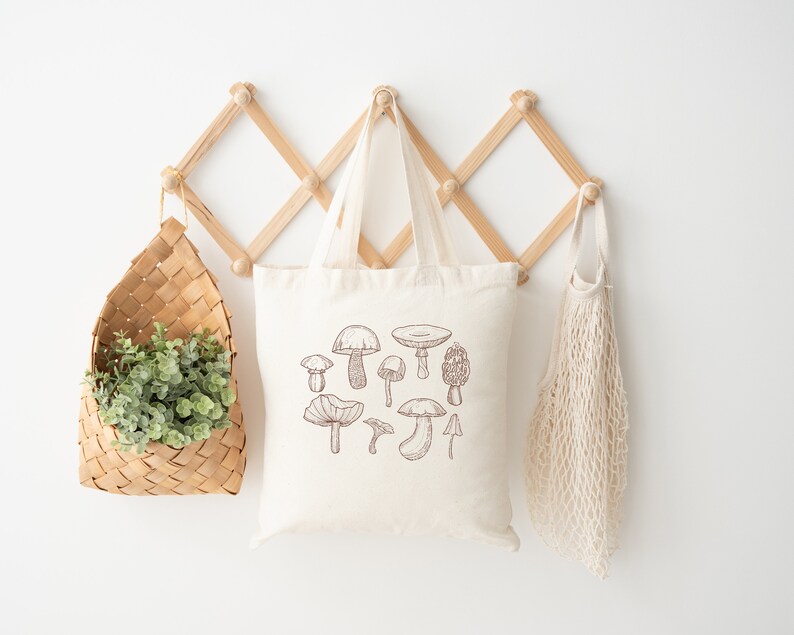 May include: A white tote bag with a brown line drawing of various mushrooms. The bag is hanging on a wooden rack with a woven basket and a mesh bag.